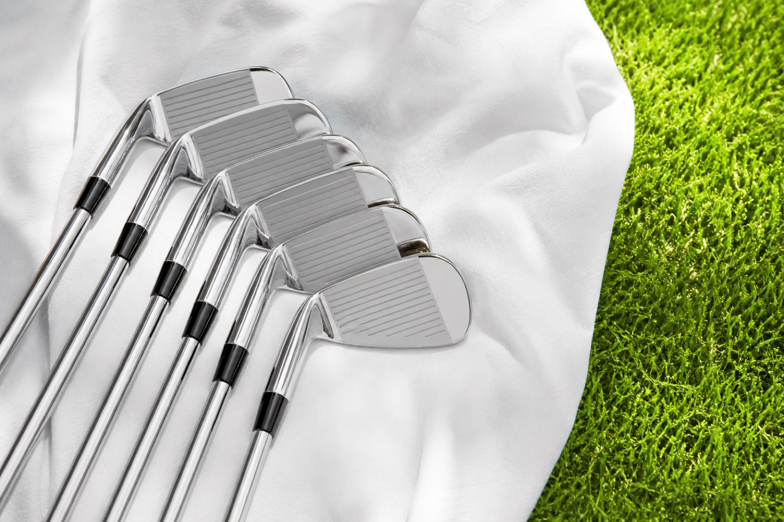 Freshly polished golf irons on a white cloth beside green grass